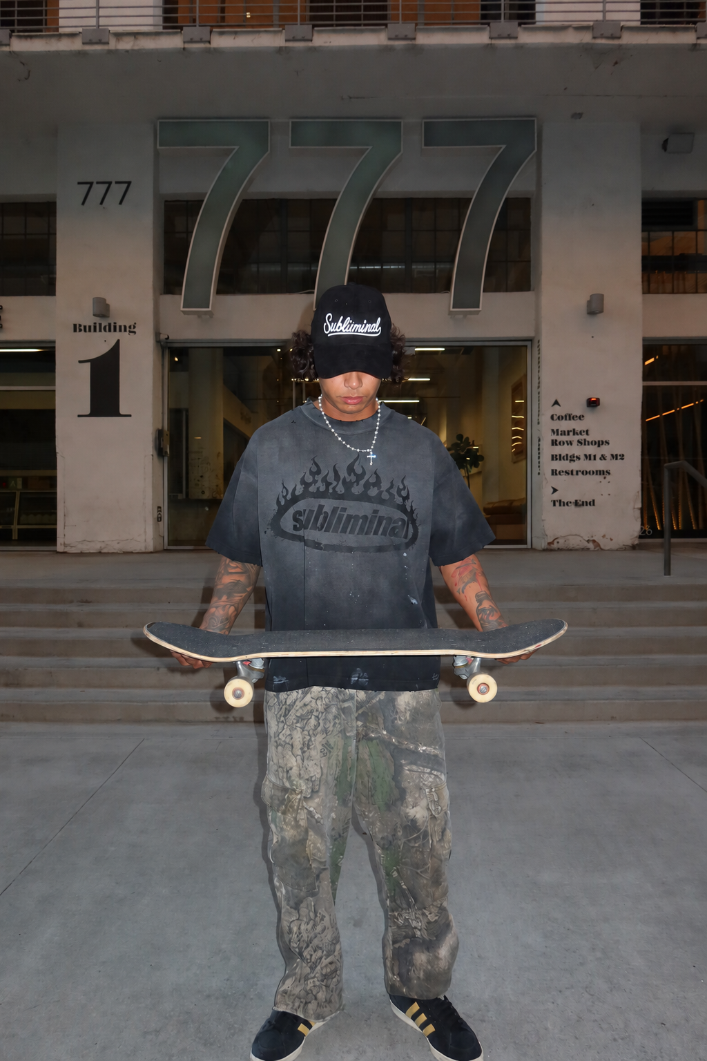 Black Logo — Distressed Tee — Black, model standing and holding a skateboard in front of the 777 building